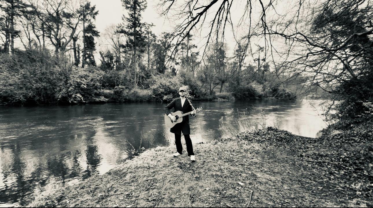 b/w picture of eoin with guitar facing camera standing beside a river